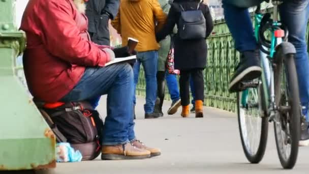 Budapest, Ungern-December 6, 2018: en hungrig tiggare sitter på asfalten av bron, har en allmosor kopp, och läser bok, människor förbi, en förbipasserande cyklist ger allm — Stockvideo