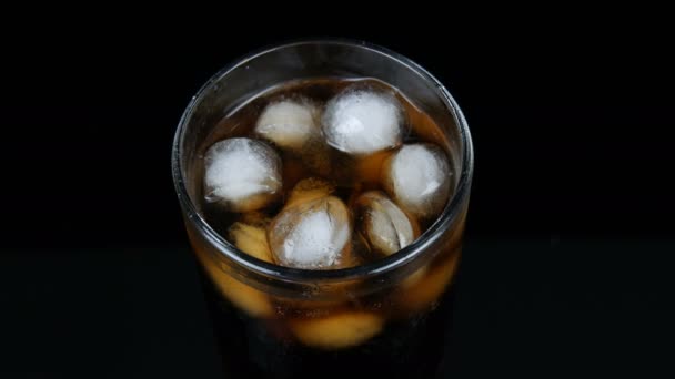 Bubbles of cold cola in a transparent long glass with ice on a black background close up view. — Stock Video