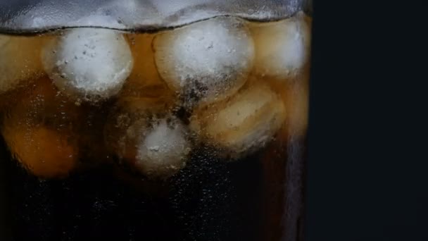 La glace est mélangée dans un verre avec des bulles de cola froid dans un long verre transparent avec de la glace sur un fond noir gros plan . — Video