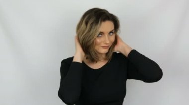 Saç dokunur siyah bir elbise güzel bir genç seksi zarif kadının portresi, saç düzeltir ve beyaz bir arka planda gülümsüyor iken kameraya bakıyor.