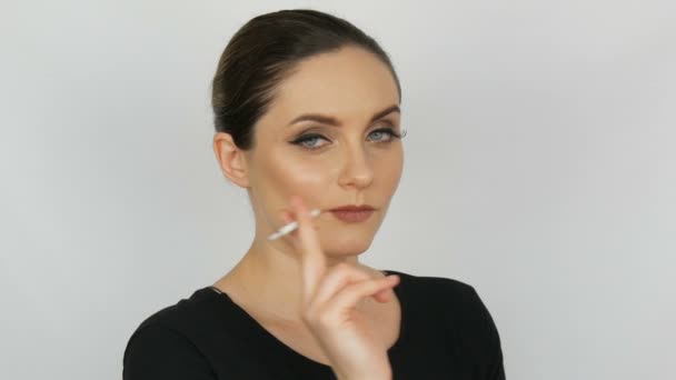 Portrét krásné elegantní mladé ženy v černých šatech s hladkým bunned vlasy a klasický make-up s velkými černými šipkami, kouřil cigaretu ladys tenké na bílém pozadí. — Stock video