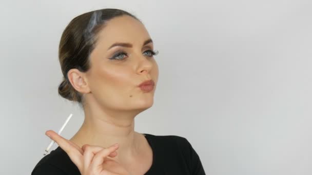 Portret van een mooie elegante jonge vrouw in een zwarte jurk met gladde bunned haar en klassieke make-up met grote zwarte pijlen, roken van een sigaret ladys dunne op een witte achtergrond. — Stockvideo