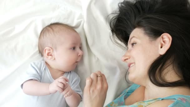 Een mooie jonge moeder en grappige twee maanden durende pasgeboren baby zijn liggend op het bed en op zoek naar elkaar. Moeder lijnen zachtjes haar baby. — Stockvideo