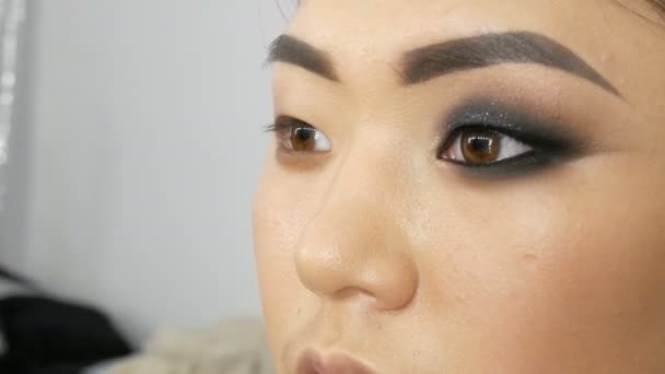Profesional estilista maquillaje artista compone el ojo humeante de una chica asiática modelo cara en visage studio — Vídeos de Stock