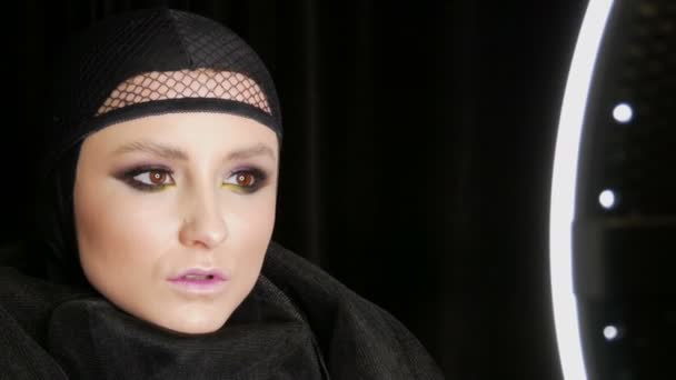 Professionell tjej modell med vacker makeup utgör i en svart mössa på huvudet framför kameran på svart bakgrund i bilden av en svart änka. Hög modegrad — Stockvideo