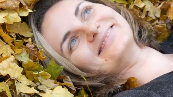 Портрет лица красивой молодой девушки без макияжа, которая лежит в осенней желтой листве и наслаждается жизнью — стоковое видео