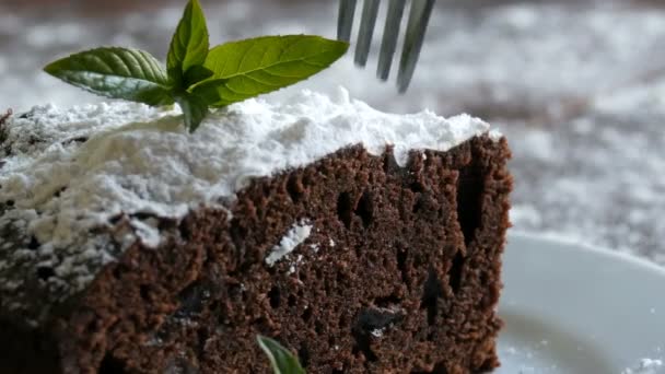 Pastel de chocolate horneado casero de brownie amortiguado con azúcar en polvo en un plato blanco decorado con hojas de menta. Tenedor rompe pedazo de pastel de brownie de la placa — Vídeo de stock