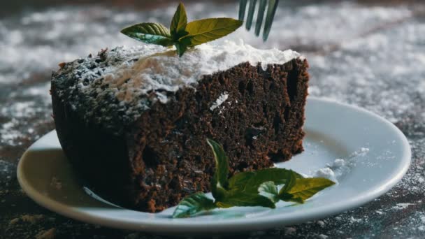 Pastel de chocolate horneado casero de brownie amortiguado con azúcar en polvo en un plato blanco decorado con hojas de menta. Tenedor rompe pedazo de pastel de brownie de la placa — Vídeo de stock