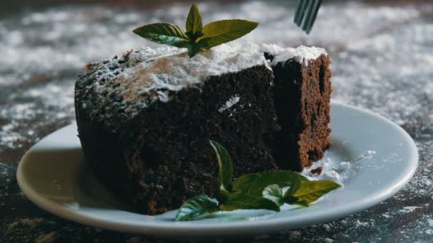 Σπιτικό φούρνο brownie σοκολάτας κέικ βουβός με ζάχαρη άχνη σε ένα λευκό πιάτο διακοσμημένο με φύλλα δυόσμου. Πιρούνι διακόπτει κομμάτι της πίτας νεράιδα από την πλάκα — Αρχείο Βίντεο