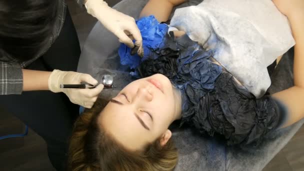 Στούντιο μόδας, το μοντέλο ετοιμάζεται για την παράσταση. Teen κορίτσι βρίσκεται σε ένα ειδικό καναπέ, σχεδιαστής προετοιμάζει την εικόνα της χρησιμοποιώντας ένα ειδικό σπρέι αερογράφου με μπογιά — Αρχείο Βίντεο