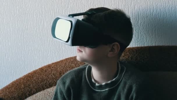 Хлопчик-підліток в білих окулярах віртуальної реальності або vr на голові сидить на дивані в кімнаті і грає . — стокове відео