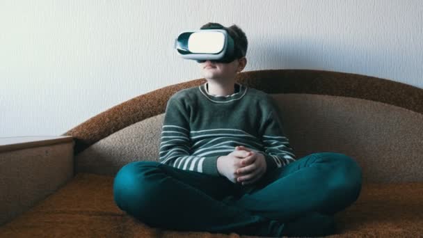 Adolescente niño en gafas blancas de realidad virtual o vr en su cabeza sentado en el sofá en la habitación y jugando . — Vídeo de stock