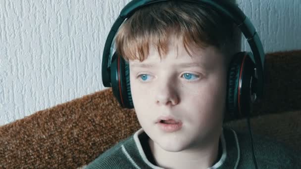 Teen boy with big headphones on his head is listening to music on a smartphone while sitting on a brown sofa. — Stock Video