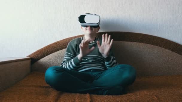 Tini fiú fehér pohár virtuális valóság vagy VR a fejét ül a kanapén a szobában és a játék. — Stock videók