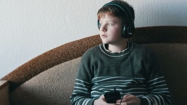 Teen boy z dużymi słuchawkami na głowie jest słuchanie muzyki na smartfonie, siedząc na brązowej kanapie. — Wideo stockowe