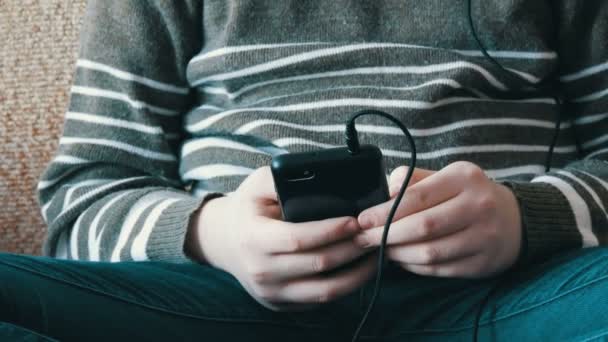 Tangan seorang remaja yang memegang smartphone hitam dan mendengarkan musik di headphone saat duduk di sofa . — Stok Video