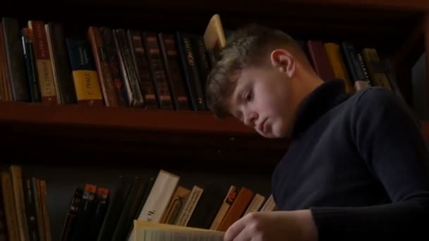 Мальчик-подросток читает книгу в библиотеке на фоне книжных полок с большим количеством книг . — стоковое видео