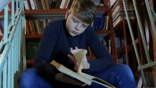 Teen boy czyta książkę w bibliotece na tle regałów z wieloma książkami. — Wideo stockowe