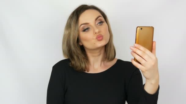 Portret van een mooie jonge sexy elegante vrouw in een zwarte jurk die neemt een selfie op een smartphone en bewonder zichzelf tegen een witte achtergrond — Stockvideo