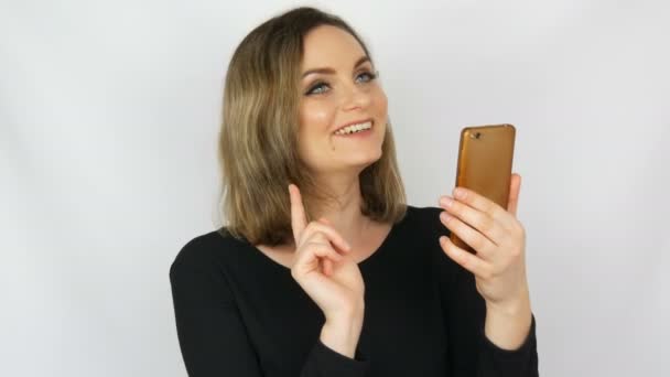 Retrato de una hermosa joven sexy y elegante mujer en un vestido negro que toma una selfie en un teléfono inteligente y se admira sobre un fondo blanco — Vídeos de Stock