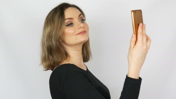 Retrato de una hermosa joven sexy y elegante mujer en un vestido negro que toma una selfie en un teléfono inteligente y se admira sobre un fondo blanco — Vídeos de Stock