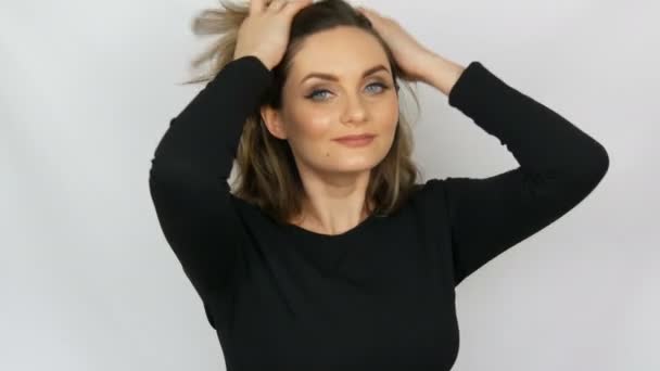 Portrét krásné mladé sexy elegantní ženy v černých šatech, které se jí dotýkala vlasů, narovnala vlasy a dívá se do kamery při úsměvu na bílém pozadí. — Stock video