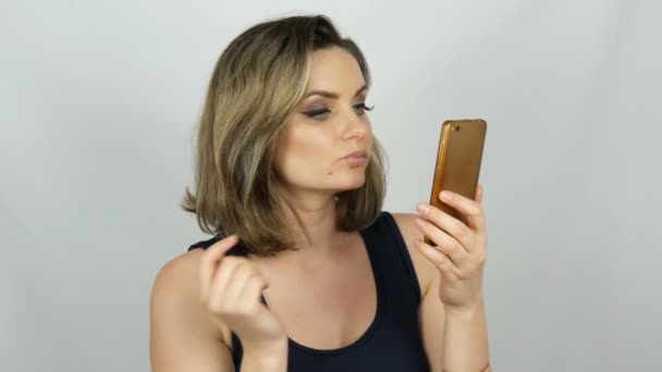 Retrato de uma bela jovem que está posando fazendo uma selfie e se comunicando e olhando para um smartphone que está segurando em suas mãos no fundo branco no estúdio — Vídeo de Stock