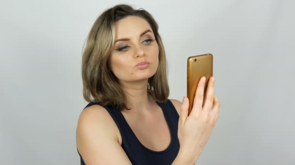 Портрет красивой молодой женщины, которая позирует, делая селфи и общаясь и глядя в смартфон, который держит в руках на белом фоне в студии — стоковое видео