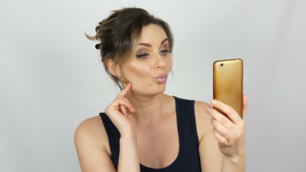 Ritratto di una bella giovane donna che posa facendo un selfie e comunicando e guardando in uno smartphone che tiene tra le mani su sfondo bianco in studio — Video Stock