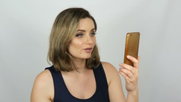 Retrato de uma bela jovem que está posando fazendo uma selfie e se comunicando e olhando para um smartphone que está segurando em suas mãos no fundo branco no estúdio — Vídeo de Stock