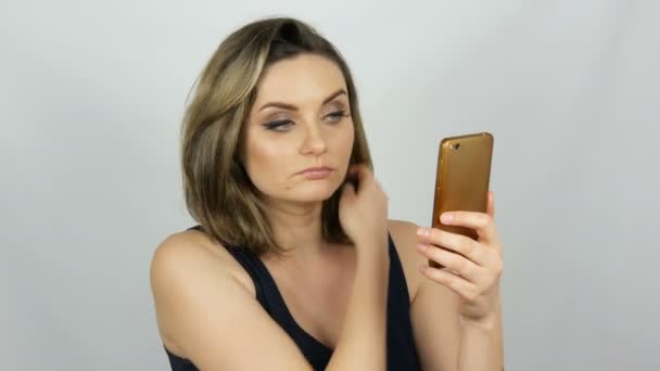 Portret van een mooie jonge vrouw die poseert het maken van een selfie en communiceren en op zoek naar een smartphone die in haar handen houdt op witte achtergrond in de Studio — Stockvideo