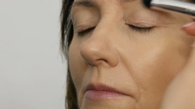 Sanatçı makyaj mavi gözlü orta yaşlı kadına vurgulayıcı kapatıcı ile makyaj yapmak. Yaş makyajı. Yakın çekim portre. Fırça ile tozu Uygula