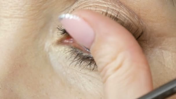 Профессиональный визажист приклеивает накладные ресницы на голубые глаза взрослой женщины средних лет вблизи. — стоковое видео