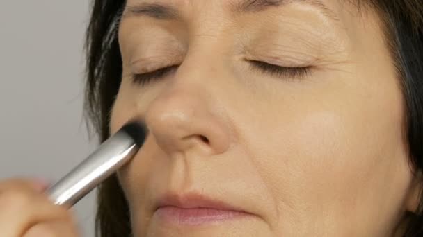 Maquillaje artista hacer maquillaje con corrector corrector a la mujer de mediana edad con ojos azules. Maquillaje de edad. Retrato de cerca. Aplicar polvo con pincel — Vídeos de Stock