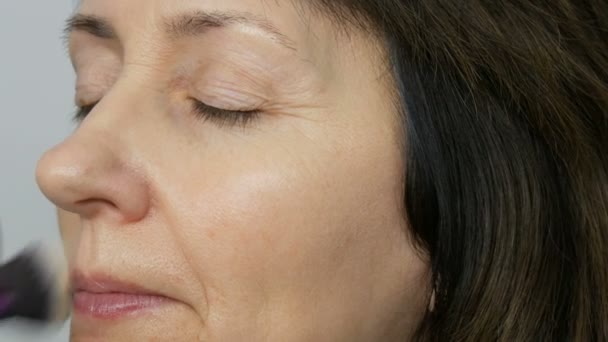 Make up καλλιτέχνη κάνει μακιγιάζ με το κονσίλερ επισήμανσης σε μεσήλικες γυναίκα με μπλε μάτια. Μακιγιάζ ηλικίας. Κοντινό πορτρέτο. Εφαρμόστε σκόνη με βούρτσα — Αρχείο Βίντεο