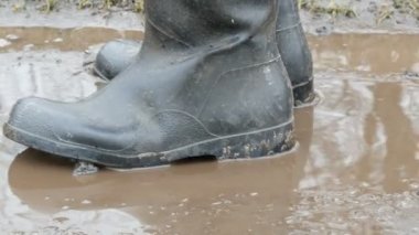 Bir adam kauçuk botlarda çamurlu su birikintisi boyunca yürür.