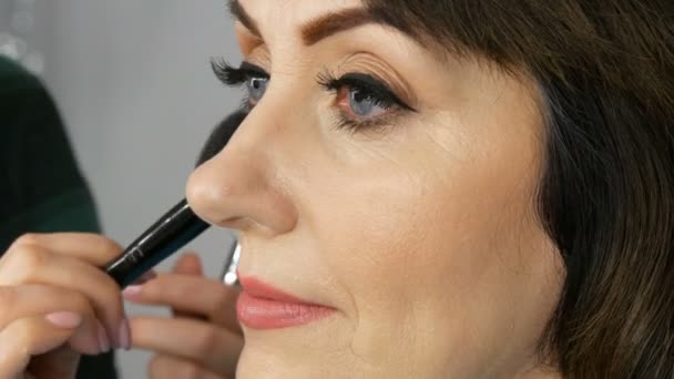 Professionele make-up Artist maakt make-up mooie middelbare leeftijd volwassen vrouw met grote blauwe ogen en schilderde de pijlen. Speciale borstel poeder gezicht — Stockvideo