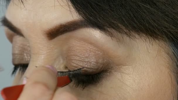 Artista de maquillaje profesional pega pestañas falsas en los ojos azules de una mujer adulta de mediana edad vista de cerca — Vídeos de Stock