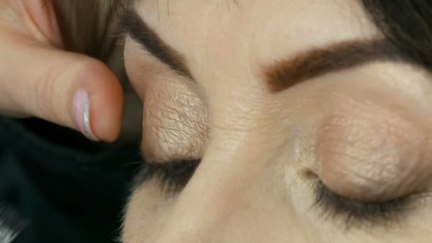 Artista de maquiagem profissional começa a colar pestanas falsas nos olhos azuis de uma mulher adulta de meia-idade vista de perto — Vídeo de Stock
