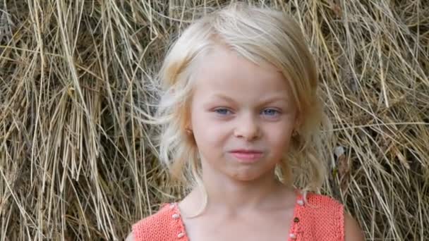 干し草の山を背景に市外の通りで汚れた顔の子供を持つかわいい笑顔と笑う青い目のブロンドの7歳の女の子の肖像画 — ストック動画