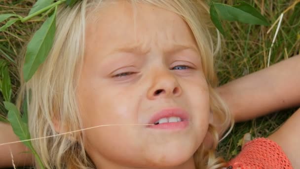 Linda rubia de ojos azules niña de siete años con una cara sucia. La niña está acostada en la hierba con las manos detrás de la cabeza masticando paja seca. Cara de cerca — Vídeos de Stock