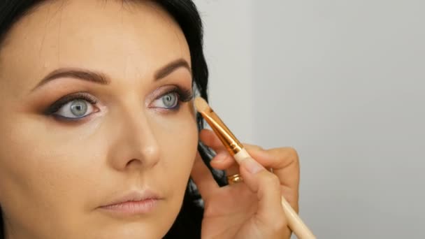 Retrato de uma bela modelo de mulher de olhos azuis com cabelo preto longo que está fazendo maquiagem olho marrom à noite e sobrancelha com escova especial — Vídeo de Stock