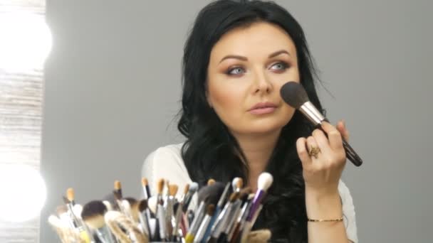 Porträt einer schönen blauäugigen Frau mit langen schwarzen Haaren, die vor dem Spiegel sitzt und ihr Make-up mit einem speziellen Puderpinsel einstellt — Stockvideo