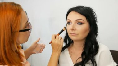 Kırmızı saçlı stilist makyaj sanatçısı güzellik salonunda uzun siyah saçlı model şık akşam makyaj yapıyor