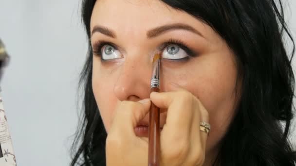 Retrato de una hermosa modelo de mujer de ojos azules con el pelo largo y negro que está haciendo maquillaje de ojos marrón noche y ceja con cepillo especial — Vídeos de Stock