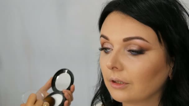 Pelirroja estilista maquillaje artista haciendo elegante noche maquillaje de modelo con pelo largo negro en salón de belleza — Vídeo de stock