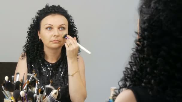 Mulher morena nova bonita com cabelo preto encaracolado faz-se maquiagem na frente do espelho — Vídeo de Stock