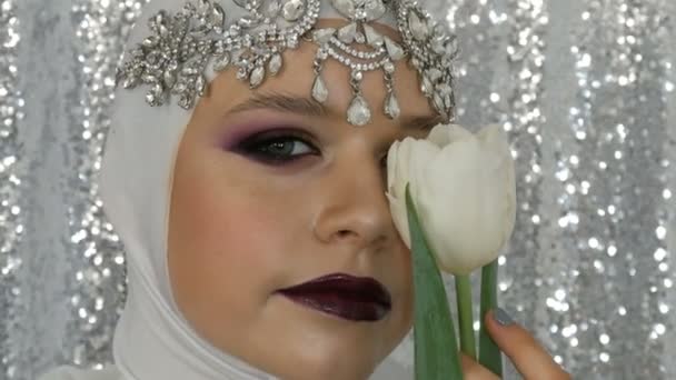 Cara de modelo y tulipán en una imagen blanca y una tiara plateada con maquillaje multicolor brillante llamado ojo humeante, posando frente a la cámara en agencia de modelos sobre un fondo plateado. Alta moda — Vídeos de Stock