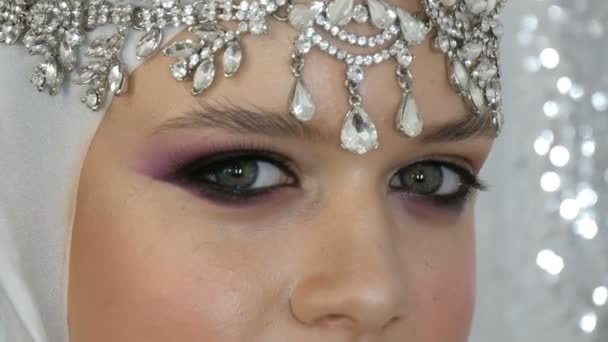 Face of model in a white image and a silver tiara with bright multi-colored make-up called a smoky eye, posing in front of the camera in model agency on a silver background. High fashion — Stock Video