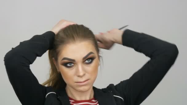 Ein schönes junges Model mit einem modischen Make-up namens Smoky Eye steht vor einem Spiegel und lässt sich die Haare glätten. — Stockvideo